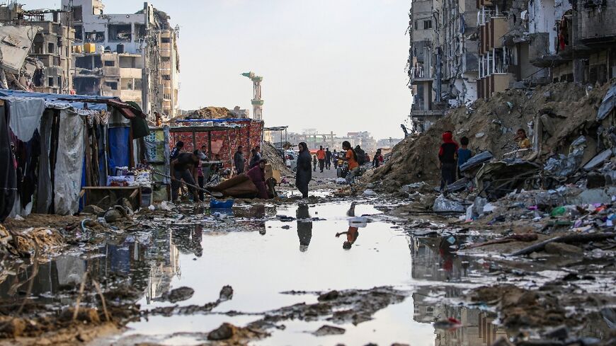 Displaced Palestinians walk around a puddle in front of destroyed buildings and tents in Khan Yunis