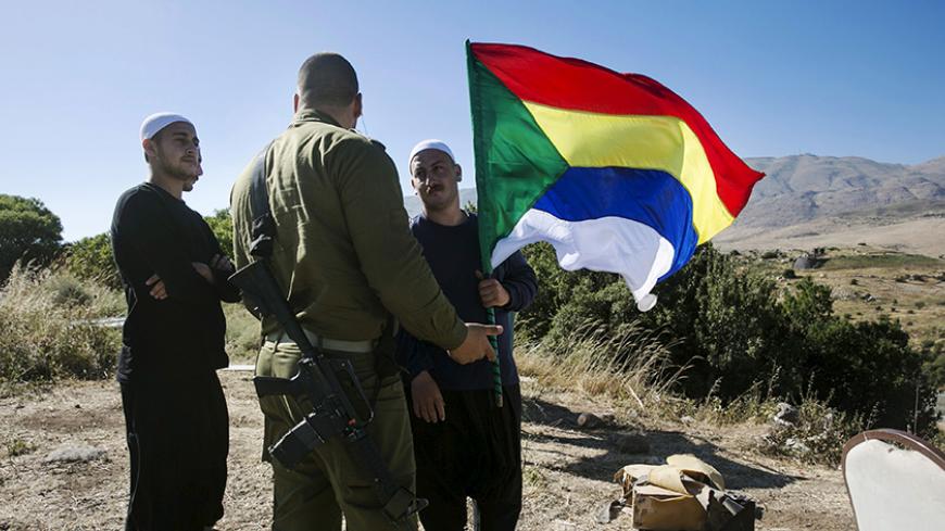 A member of the Druze community holds a Druze flag as he speaks to an Israeli soldier near the border fence between Syria and the Israeli-occupied Golan Heights, near Majdal Shams June 18, 2015. Gathered at a hilltop in the Israeli-held Golan Heights, a group of Druze sheikhs look through binoculars at the Syrian village of Hader, surrounded by Islamist fighters. They fear their brethren are in imminent danger. Intense battles this week have left Syria's al Qaeda branch, the Nusra Front, in control of hills