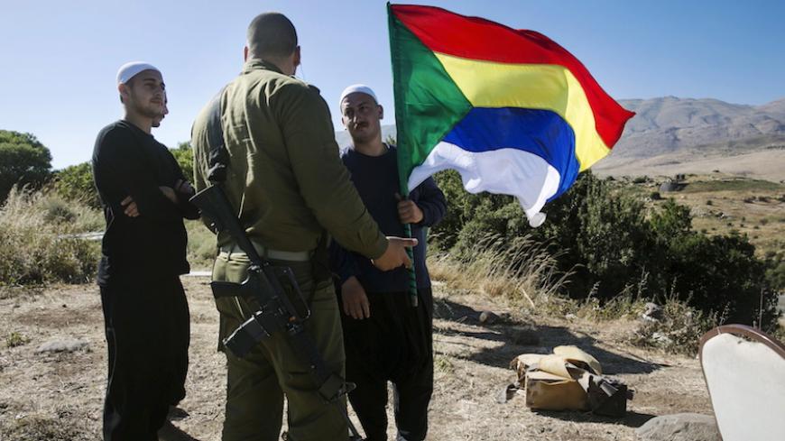 A member of the Druze community holds a Druze flag as he speaks to an Israeli soldier near the border fence between Syria and the Israeli-occupied Golan Heights, near Majdal Shams June 18, 2015. Gathered at a hilltop in the Israeli-held Golan Heights, a group of Druze sheikhs look through binoculars at the Syrian village of Hader, surrounded by Islamist fighters. They fear their brethren are in imminent danger. Intense battles this week have left Syria's al Qaeda branch, the Nusra Front, in control of hills