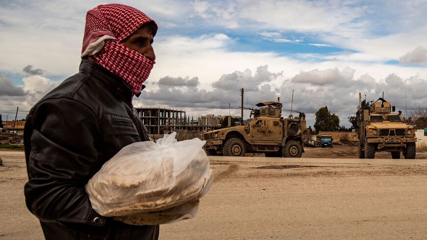 A man carrying bags of bread walks as a patrol of US military vehicles is seen in the town of Tal Tamr in the northeastern Syrian Hasakeh province along the border with Turkey on Feb. 8, 2020.