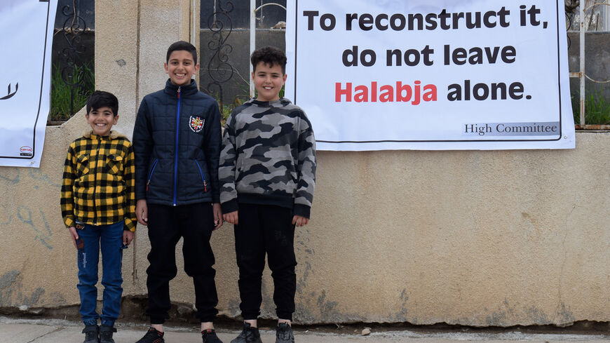 Children stand next to a sign reading "To reconstruct it, do not leave Halabja alone" by the Martyrs' Cemetery in Halabja on March 16, 2022. 