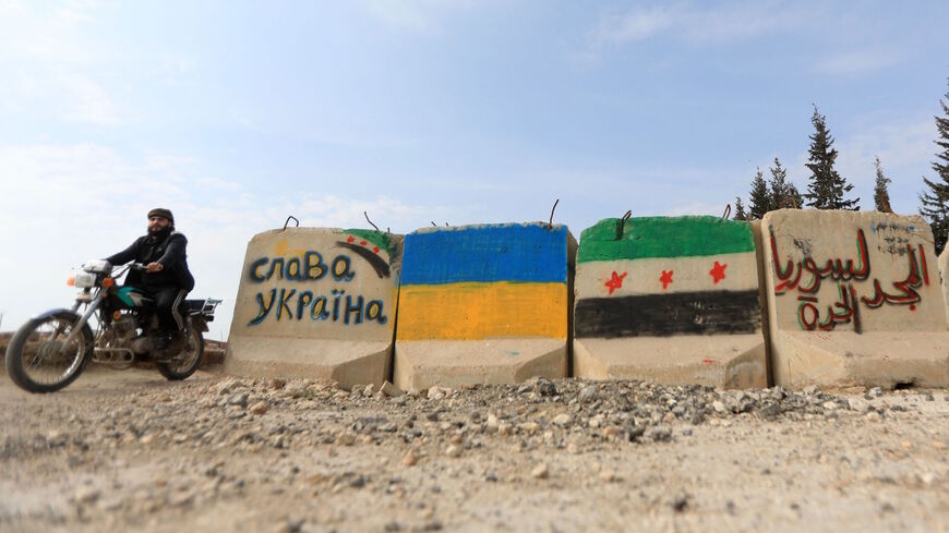 A man rides a motorcycle past cement blocks bearing flags of Ukraine (C L) and the Syrian opposition with inscriptions, one in Cyrillic (L) which reads "Slava Ukraini" ("glory to Ukraine"), a Ukrainian national salute, and another in Arabic which reads "glory to free Syria", near the Syrian rebel-held city of al-Bab in the northern Aleppo governorate, on Feb. 28, 2022.