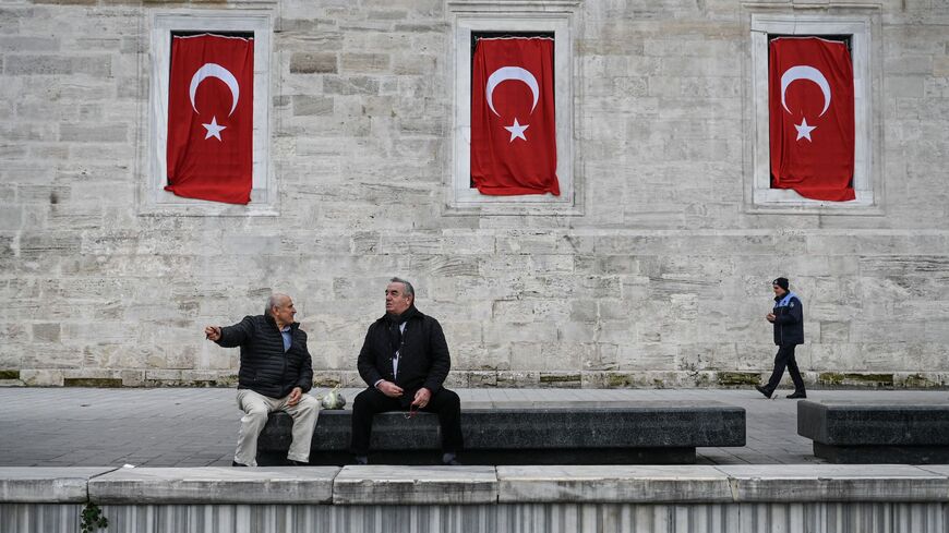 Men chat as they sit on a bench in front of the New Mosque, bearing Turkish flags, in the Eminonu district in Istanbul, on January 10, 2023. (Photo by OZAN KOSE / AFP) (Photo by OZAN KOSE/AFP via Getty Images)