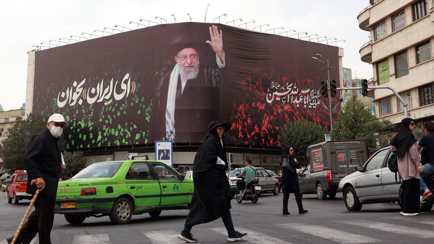 People cross a street next to a billboard bearing the portrait of Iranian Supreme Leader Ayatollah Ali Khamenei and a quote that reads "Sing Oh Iran" in Tehran on July, 9, 2025. 