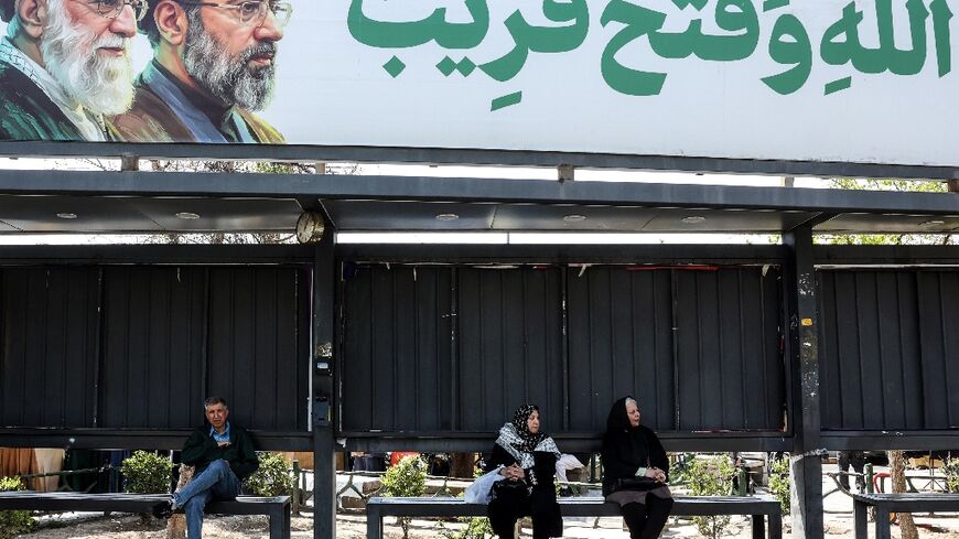 Iranians wait at a bus stop as they sit  under a banner featuring Iran's late supreme leaders Ayatollah Ali Khamenei (L) and his son newly elected supreme leader Mojtaba Khamenei, in northern Tehran