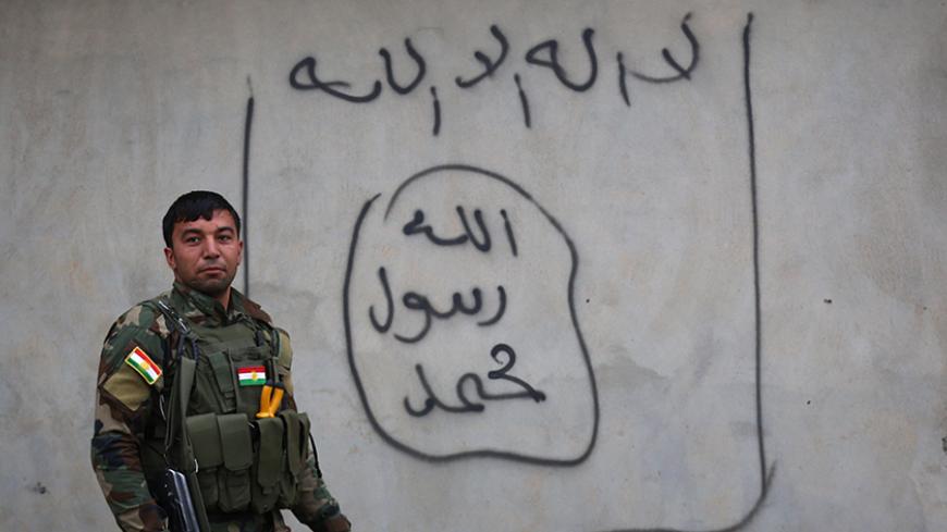A Peshmerga fighter stands next graffiti of the Islamic State (IS) group flag on a wall in Zummar city in the northern Iraqi Governorate of Nineveh on December 18, 2014, as they continue to battle Islamic State group fighters near the border with Syria. Kurdish forces backed up by US-led warplanes have recaptured a large area in Iraq near the Syrian border in an offensive this week against Islamic State jihadists, a US commander said. AFP PHOTO / SAFIN HAMED        (Photo credit should read SAFIN HAMED/AFP/