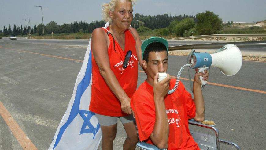 Ilana Azoulay, 53, with an Israeli flag hanging on her back, pushes her handicapped son Yossi, 17, in his wheelchair on the road to Jerusalem near Yesodot 13 July 2003. Azoulay has been pushing her handicapped teenage son in his wheelchair half the length of Israel in scorching heat -- one of hundreds of mothers converging on Jerusalem to protest against new austerity measures which deprive them of much needed state benefits. Azoulay is on a mission to make the voice of Israel's most deprived heard. Sympath