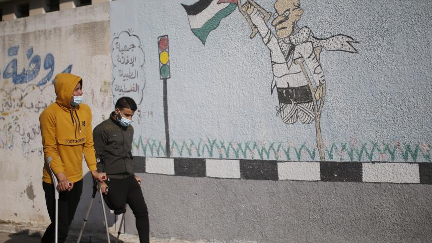 Two Palestinian youths on crutches wearing a protective mask due to the Covid-19 pandemic, walk past graffiti depicting a disabled man carrying a national flag, during an event marking the International Day of Persons with Disabilities in Gaza City on December 3, 2020. (Photo by MOHAMMED ABED / AFP) (Photo by MOHAMMED ABED/AFP via Getty Images)