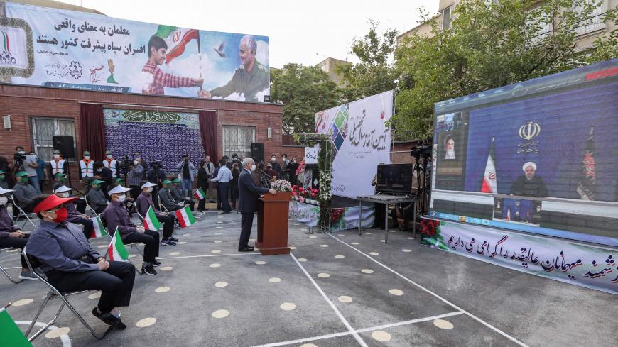 Mask-clad school children hold Iranian national flags as they sit distanced apart from each other in the yard of a school while attending a speech by Iran's President Hassan Rouhani by video conference, at Nojavanan school in the capital Tehran on the first day of schools re-opening on September 5, 2020, while in the background a billboard is seen showing a boy holding a flag together with Qasem Soleimani, the late commander of Iran's Revolutionary Guard Corps (IRGC) next to a Farsi quote by the country's s