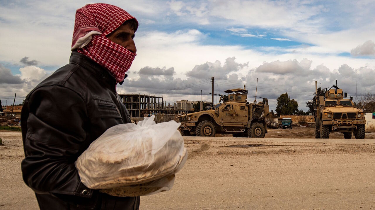 A man carrying bags of bread walks as a patrol of US military vehicles is seen in the town of Tal Tamr in the northeastern Syrian Hasakeh province along the border with Turkey on Feb. 8, 2020.