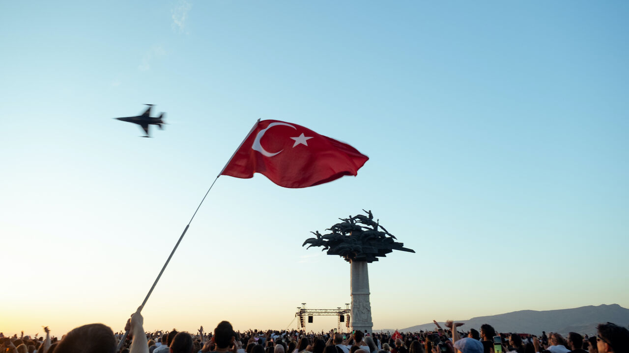Photo of a waving Turkish flag in the sky with a jet flyover behind, and a crowd below holding flags, captured on Izmir's liberation anniversary.