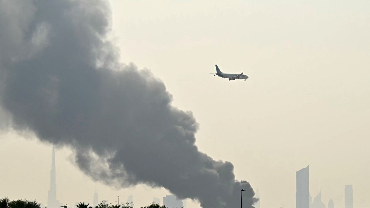 An Emirates aircraft flies past plumes of smoke from an ongoing fire near Dubai International Airport, Dubai, March 16, 2026.