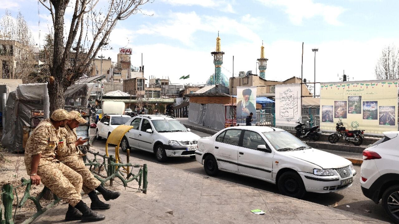 Two Iranian soldiers look at their mobile phone as they sit close to an image of Iran's slain supreme leader Ayatollah Ali Khamenei (R), along a street close to Tajrish Square in Tehran on March 7, 2026