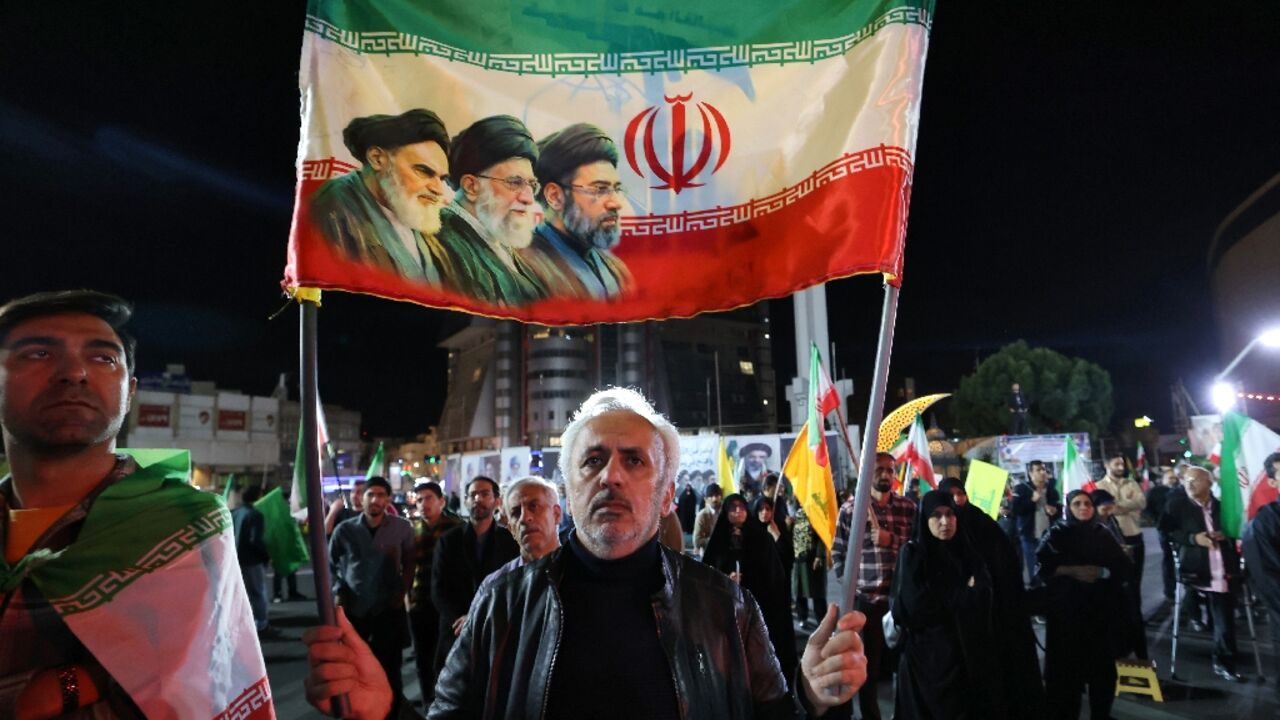 A man carries an Iranian flag with the portraits of Iran's late supreme leader, Ayatollah Ruhollah Khomeini (L), slain supreme leader Ayatollah Ali Khamenei (C) and Iran's supreme leader Ayatollah Mojtaba Khamenei as people rally in his support