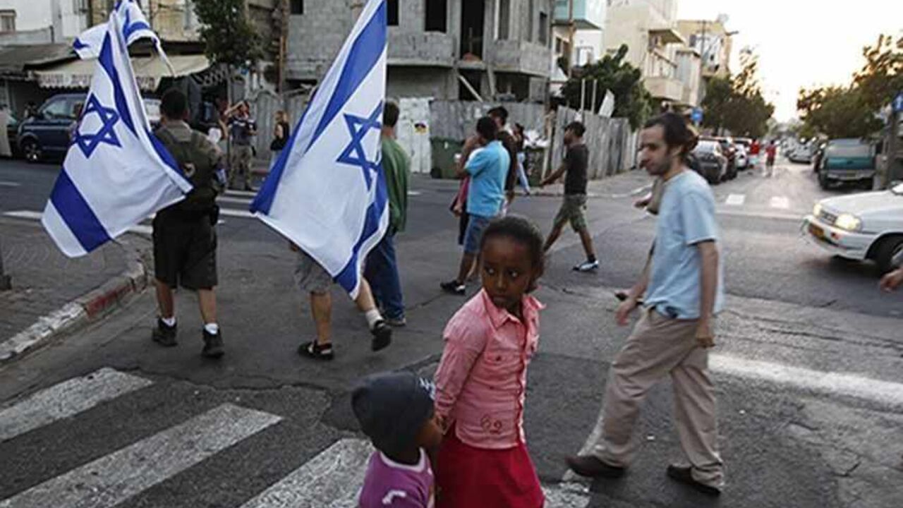 Children cross the street as residents of south Tel Aviv carry Israeli flags during a protest against African migrants living in their neighbourhoods May 30, 2012. Last week a similar protest held by residents of the low-income neighbourhoods where many of the border-jumpers from Eritrea, Sudan and South Sudan live led to violence, including a rampage that an Israeli broadcaster dubbed a "pogrom". Fleeing poverty, fighting and authoritarian rule, some 60, 000 Africans have crossed illegally into Israel thro