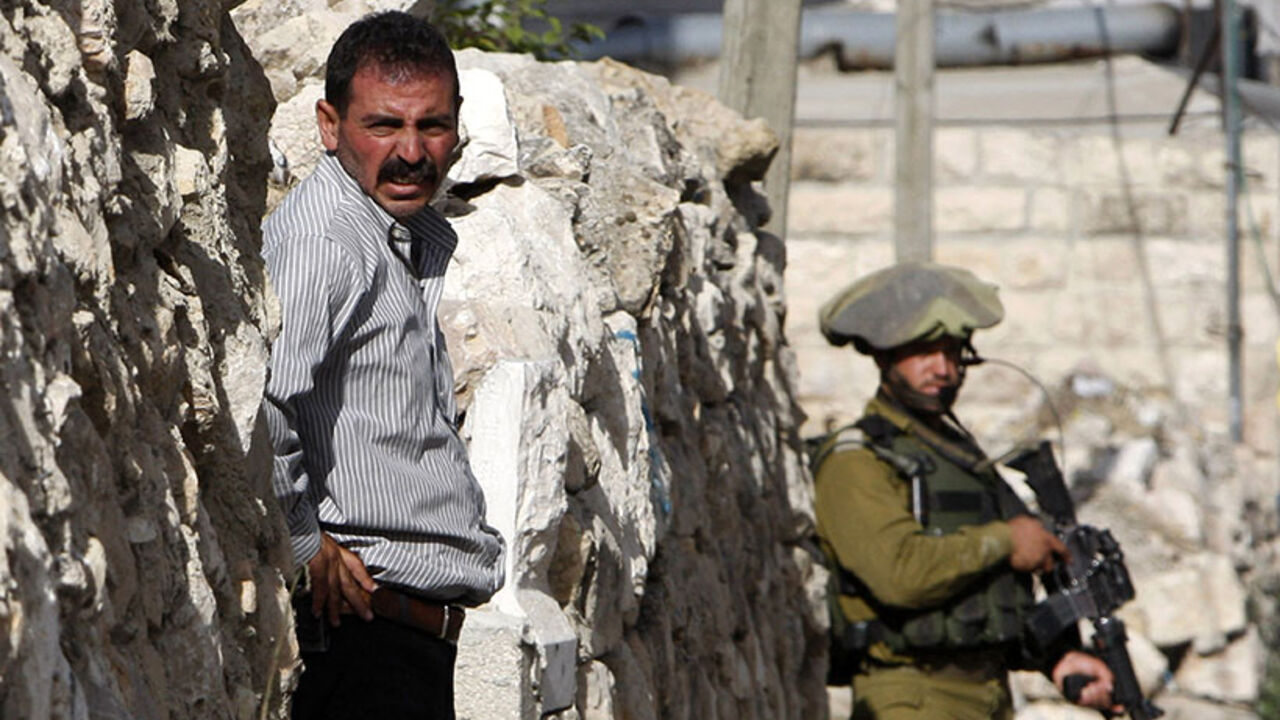 A Palestinian man stands outside his house as an Israeli soldier takes part in an operation to locate three Israeli teens in the West Bank City of Hebron June 19, 2014. Israeli forces traded gunfire with Palestinians on Thursday, the military said, in the fiercest street battles in the occupied West Bank since a search began for three Israeli teenagers missing for a week. REUTERS/Mussa Qawasma (WEST BANK - Tags: POLITICS MILITARY) - RTR3UOFG