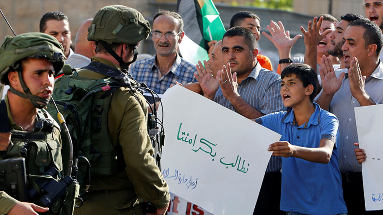 A Palestinian boy holds a sign that reads "we demand our dignity" in front of  Israeli soldiers during a protest calling for the reopening of a closed street in the West Bank city of Hebron August 24, 2016.  REUTERS/Mussa Qawasma  - RTX2MW1E