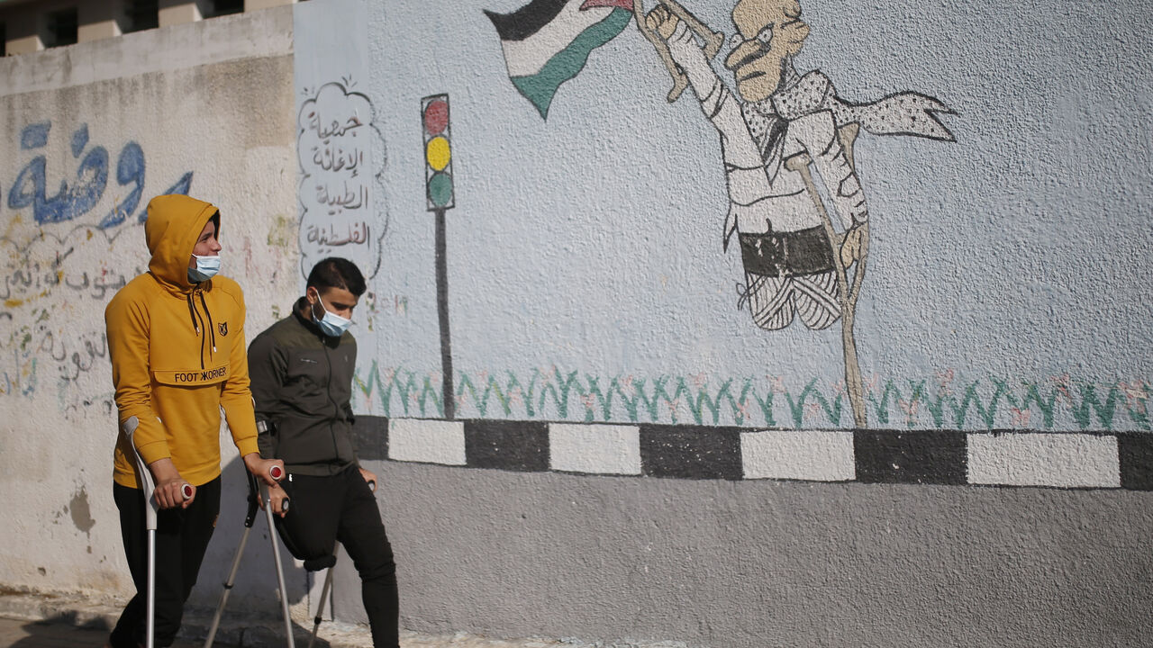 Two Palestinian youths on crutches wearing a protective mask due to the Covid-19 pandemic, walk past graffiti depicting a disabled man carrying a national flag, during an event marking the International Day of Persons with Disabilities in Gaza City on December 3, 2020. (Photo by MOHAMMED ABED / AFP) (Photo by MOHAMMED ABED/AFP via Getty Images)