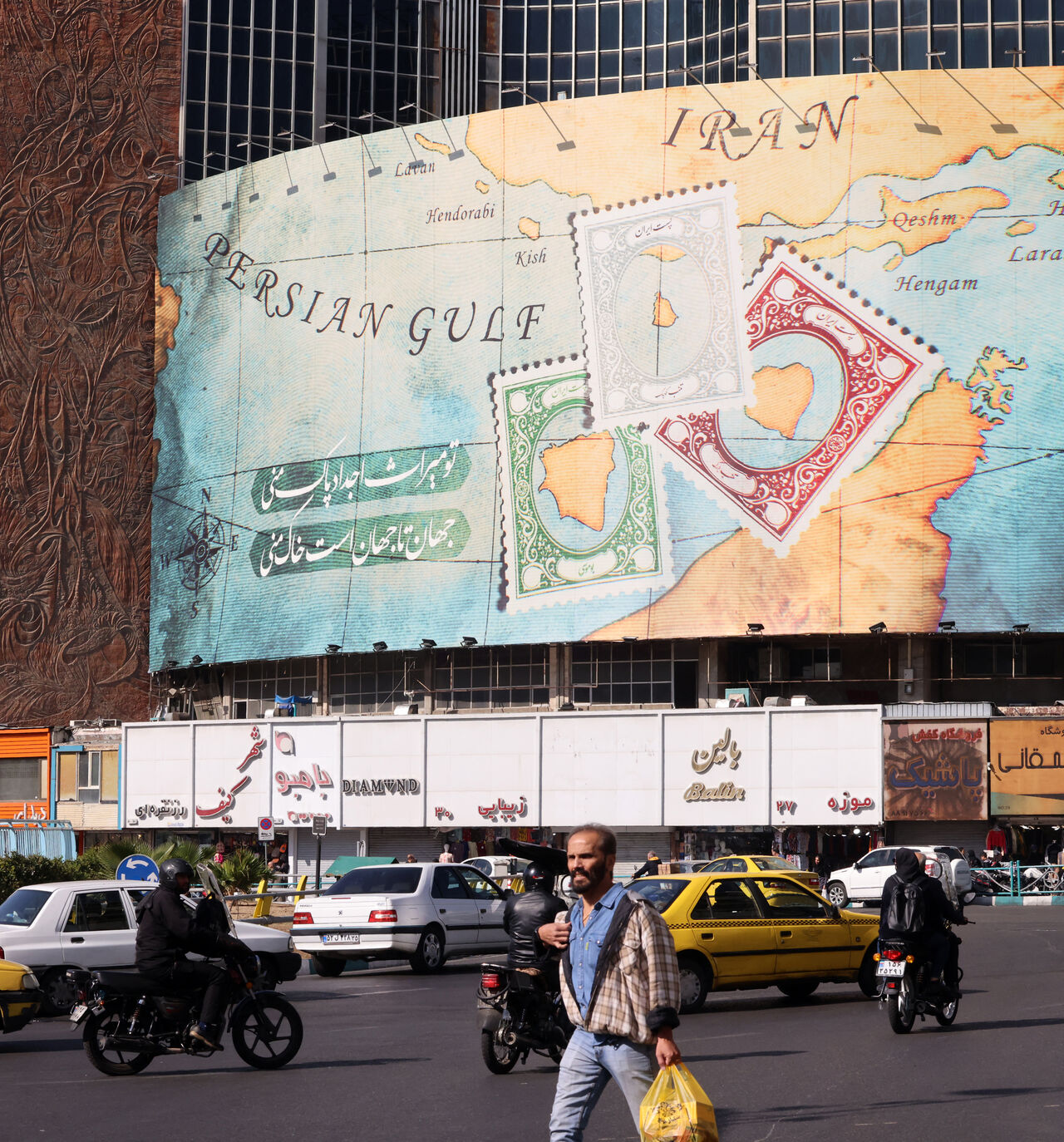 People walk and drive past a billboard covering the facade of a building on Vali-Asr square, depicting as postage stamps the disputed Abu Musa and Greater and Lesser Tunb islands that were seized by Iran in 1971, in Tehran on October 26, 2024. Residents of Tehran awoke and went about their business as planned on October 26 after their sleep was troubled by Israeli strikes that triggered blasts that echoed across the city. (Photo by ATTA KENARE / AFP) (Photo by ATTA KENARE/AFP via Getty Images)