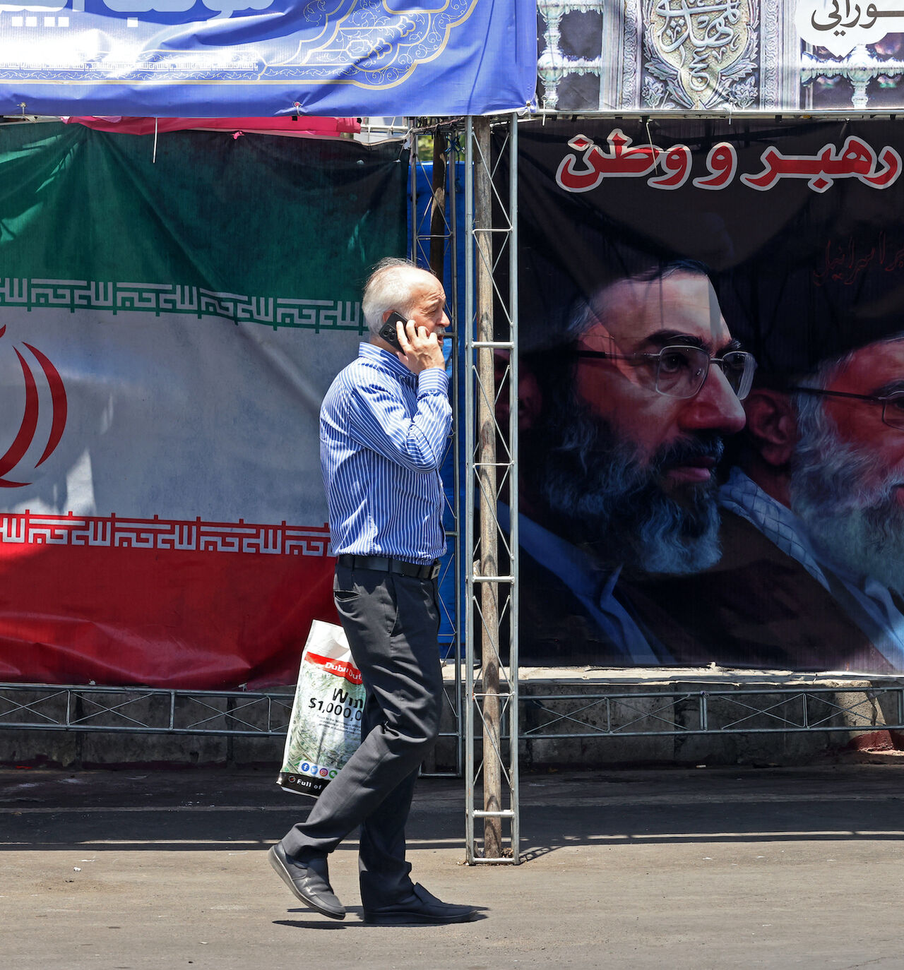 A man walks past Iran's national flag and a banner bearing the images of the late founder of the Islamic Revolution supreme leader Ayatollah Ruhollah Khomeini (R), the late Iranian supreme leader Ali Khamenei and his son, the current supreme leader Mojtaba Khamenei (L), erected along a street at the Vanak Square in Tehran on June 10, 2026.