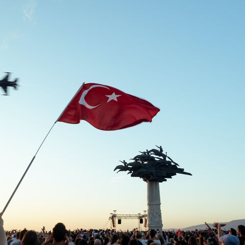 Photo of a waving Turkish flag in the sky with a jet flyover behind, and a crowd below holding flags, captured on Izmir's liberation anniversary.