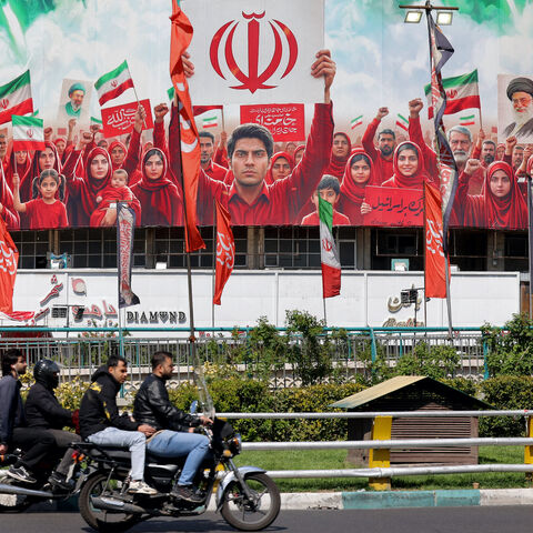 Motorists ride scooters near a large political banner, depicting missiles being fired behind Iranian demonstrators in solidarity with the government, at Valiasr Square in Tehran on April 6, 2026. 