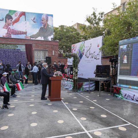 Mask-clad school children hold Iranian national flags as they sit distanced apart from each other in the yard of a school while attending a speech by Iran's President Hassan Rouhani by video conference, at Nojavanan school in the capital Tehran on the first day of schools re-opening on September 5, 2020, while in the background a billboard is seen showing a boy holding a flag together with Qasem Soleimani, the late commander of Iran's Revolutionary Guard Corps (IRGC) next to a Farsi quote by the country's s