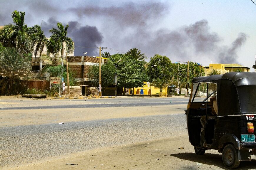 Smoke rises from buildings in Khartoum as a tuktuk taxi driver sits in his vehicle along a deserted street: most families are sheltering at home too scared to move