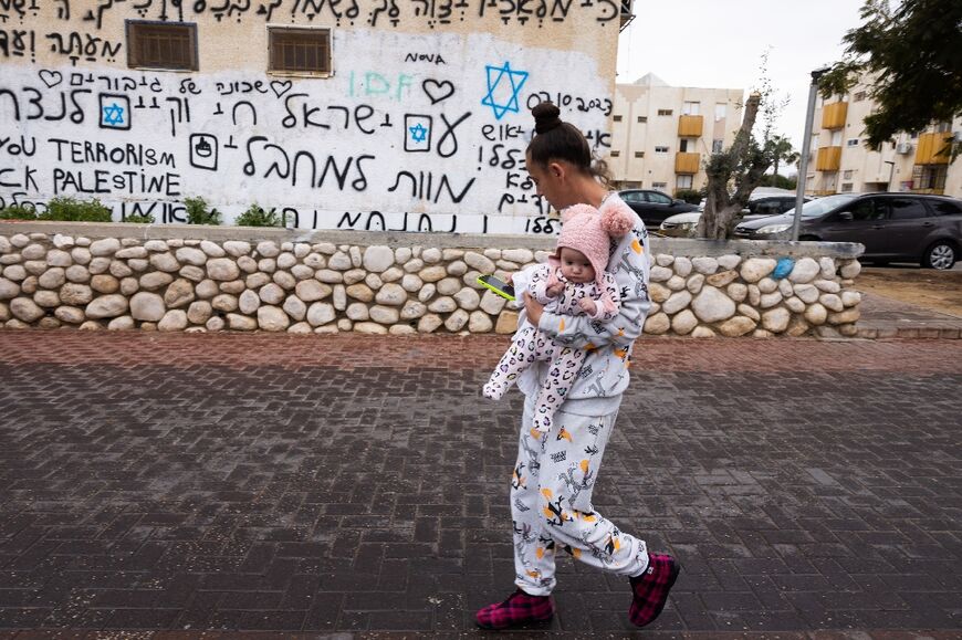 An Israeli woman carries a baby past graffiti in Hebrew and English with slogans against Hamas and condemning the October 7 attack 