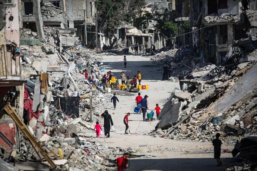 Displaced Palestinians walk along a devastated street as others gather at a water point to fill their jerricans in Khan Yunis in the southern Gaza Strip on May 16, 2024