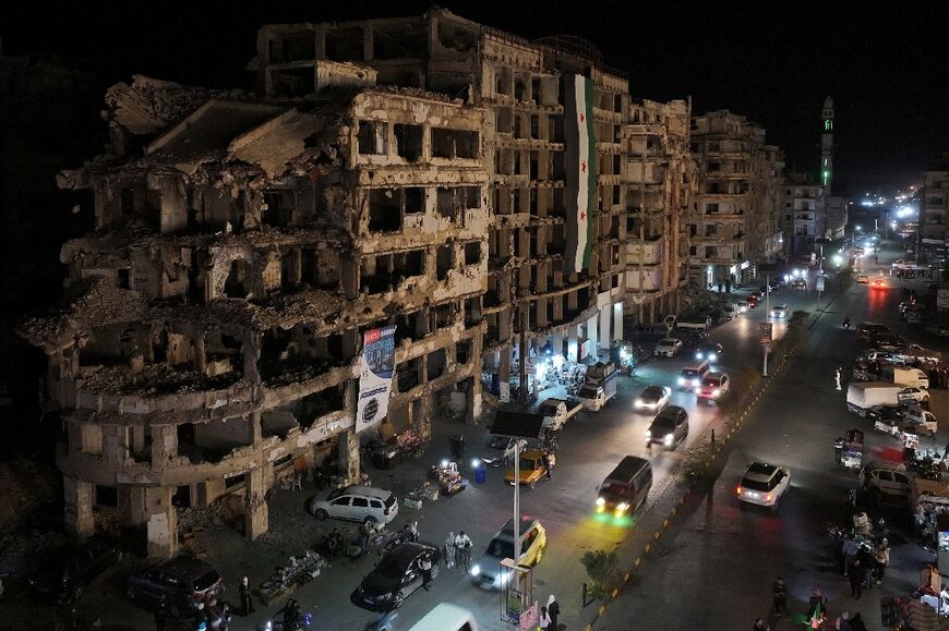 Commuters drive past damaged buildings as their city commemorates the 14th anniversary of the uprising against Syria's former president Bashar al-Assad in Homs, late on March 15, 2025