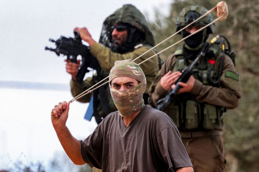 Israeli army soldiers stand behind a masked man swinging a slingshot during the incident