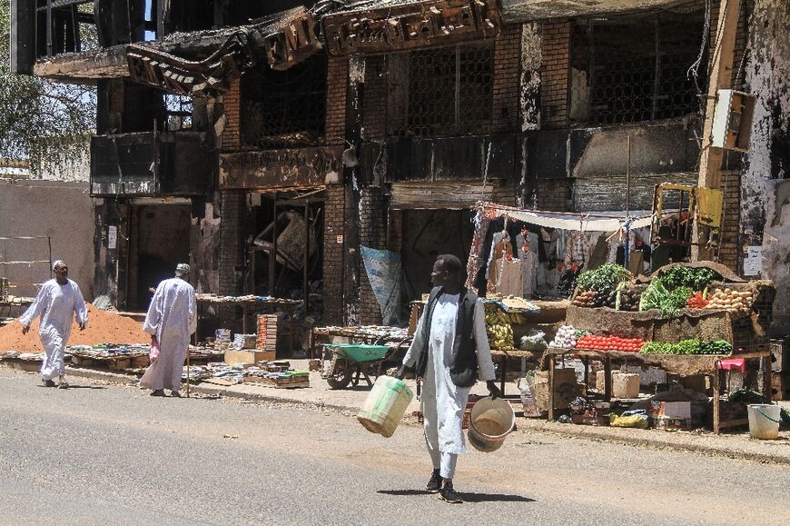 Men walk past street vendors' stands set up beneath a damaged building in the capital Khartoum on the third anniversary of the start of the war 