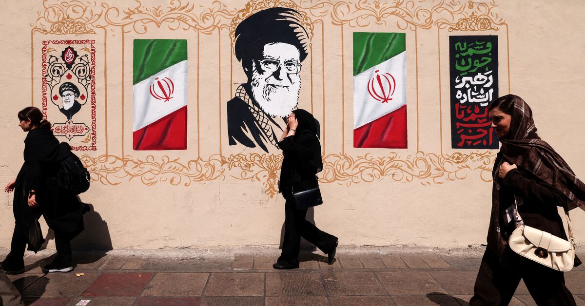 ایرانیان در تاریخ ۱۴ سپتامبر ۲۰۲۶ از کنار دیواری که با تصویر آیت‌الله علی خامنه‌ای، رهبر فقید عالی ایران و پرچم ملی تزئین شده، در مرکز تهران عبور می‌کنند. — ATTA KENARE / AFP via Getty Images