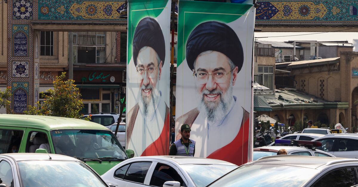 یک افسر پلیس در حال نظارت بر ترافیک است و زیر پرتره‌های رهبر عالی ایران، آیت‌الله مجتبی خامنه‌ای، در تهران ایستاده است؛ ۲۱ اکتبر ۲۰۲۶. — اتا کناره / AFP از طریق Getty Images